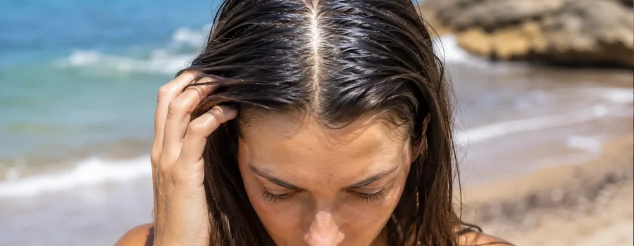 Donna al mare con capelli unti e cute oleosa durante l’estate, con radici lucide e appesantite dal sebo, sulla spiaggia davanti al mare.